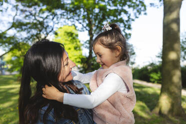 Smiling mother carrying daughter in park on sunny day - IHF00884