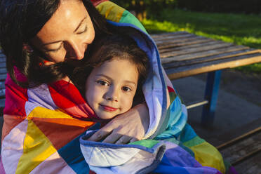 Mother and daughter sitting on bench wrapped in quilt blanket at park - IHF00903