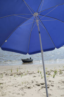 Motorboot links am Rande des Sandstrandes mit blauem Sonnenschirm im Vordergrund - GISF00868