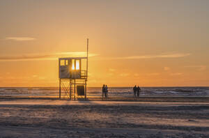 Deutschland, Niedersachsen, Juist, Rettungsschwimmerhütte bei Sonnenuntergang mit Silhouetten von Passanten im Hintergrund - KEBF02360