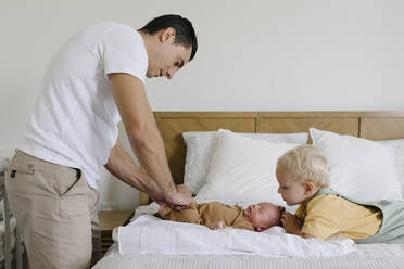 Boy looking at father buttoning baby's rompers on bed - TYF00220