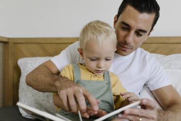 Mann liest ein Buch mit seinem Sohn, der zu Hause auf dem Bett sitzt - TYF00227