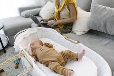 Frau mit Laptop neben dem schlafenden Baby in der Krippe zu Hause - TYF00243