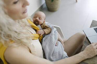 Baby boy sleeping in arms of woman working from home - TYF00248