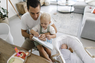 Father holding book sitting with son by baby sleeping in crib at home - TYF00253