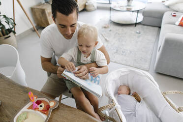 Vater hält ein Buch und sitzt mit seinem Sohn neben dem schlafenden Baby im Kinderbett - TYF00253