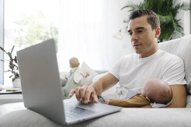 Mann mit Baby auf dem Arm und Laptop bei der Arbeit von zu Hause aus - TYF00257