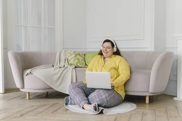 Smiling woman sitting with laptop enjoying music through wireless headphones at home - SEAF00945