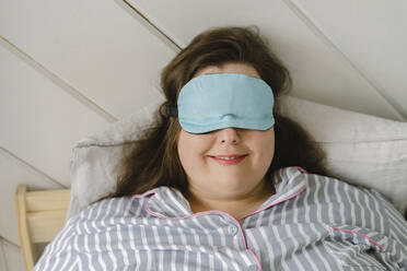 Smiling woman wearing sleeping eye mask resting on bed at home - SEAF00956