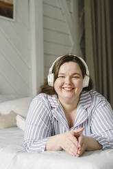 Lächelnde Frau hört Musik über drahtlose Kopfhörer auf dem Bett liegend - SEAF00959