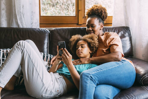 Mädchen teilt Smartphone mit Mutter auf Sofa zu Hause - MEUF06362