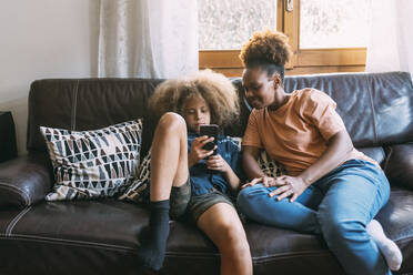 Boy using smart phone sitting with mother on sofa at home - MEUF06367