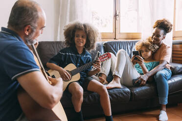 Mann, der seinem Sohn das Gitarrenspiel beibringt, sitzt mit Frau und Tochter auf dem Sofa zu Hause - MEUF06371