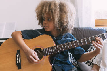 Junge mit Afrofrisur spielt Gitarre und sitzt zu Hause auf dem Sofa - MEUF06372