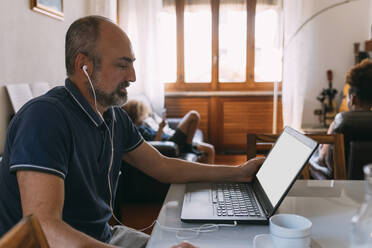 Businessman wearing in-ear headphones doing video call through laptop at home - MEUF06381