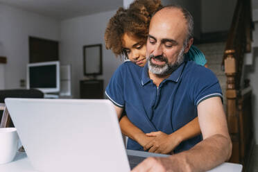 Girl embracing father using laptop working from home - MEUF06383