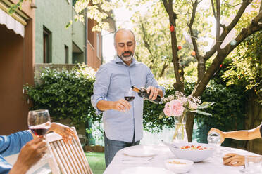 Man pouring wine into glass standing by dining table in backyard - MEUF06396