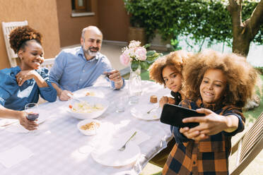 Lächelnder Junge nimmt Selfie mit Familie durch Smartphone am Esstisch im Hinterhof - MEUF06404