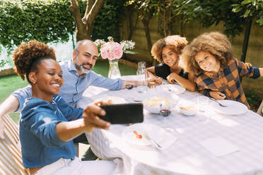 Smiling woman taking selfie with family through smart phone at dining table in backyard - MEUF06405