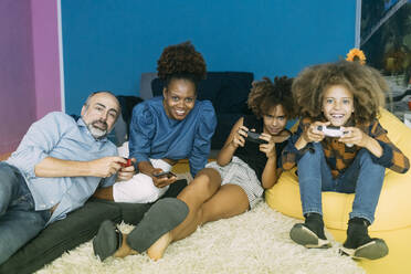 Lächelnde Frau, die ein Mobiltelefon hält, sitzt neben einem Mann und Kindern, die zu Hause ein Videospiel spielen - MEUF06416