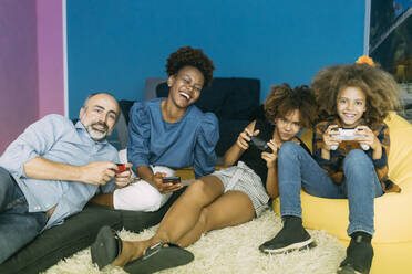 Glückliche Frau mit Handy in der Hand, die neben einem Mann und Kindern sitzt, die zu Hause ein Videospiel spielen - MEUF06417