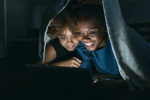 Lächelnde Frau, die ihrer Tochter zu Hause unter der Bettdecke einen Tablet-PC zeigt - MEUF06437