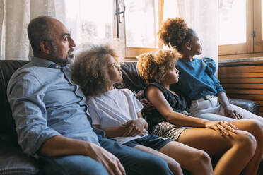 Man with family looking through window sitting on sofa at home - MEUF06443