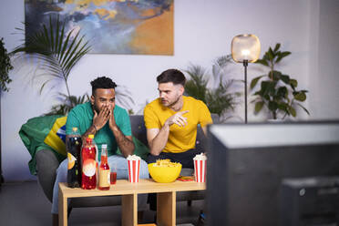 Young man talking with curious friend watching football match sitting on sofa at home - SBAF00092