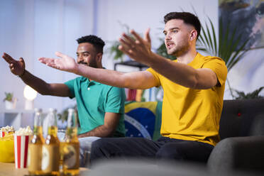Friends watching soccer match gesturing sitting on sofa in living room at home - SBAF00093