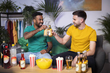 Happy young men toasting beer bottles sitting on sofa at home - SBAF00099