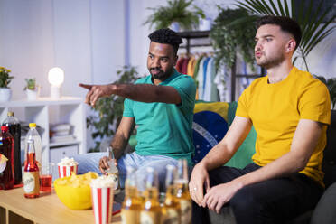 Young man with beer bottle pointing by roommate watching soccer match at home - SBAF00100