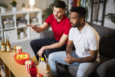 Young man talking with friend watching football match sitting on sofa at home - SBAF00113