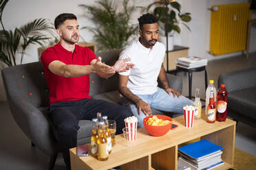 Man gesturing sitting by friend watching soccer match in living room at home - SBAF00114