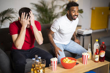 Happy man watching football match by sad roommate at home - SBAF00115