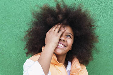 Happy girl with Afro hairstyle covering eye in front of green wall - PNAF04015