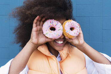 Cheerful girl covering eyes with doughnuts in front of blue wall - PNAF04048