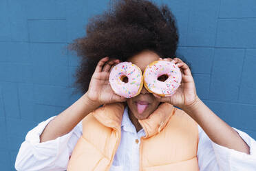 Girl sticking out tongue covering eyes with doughnuts in front of blue wall - PNAF04049