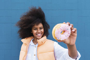Smiling girl showing doughnut standing in front of blue wall - PNAF04051