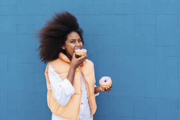 Girl with Afro hairstyle eating pink doughnuts standing by blue wall - PNAF04053