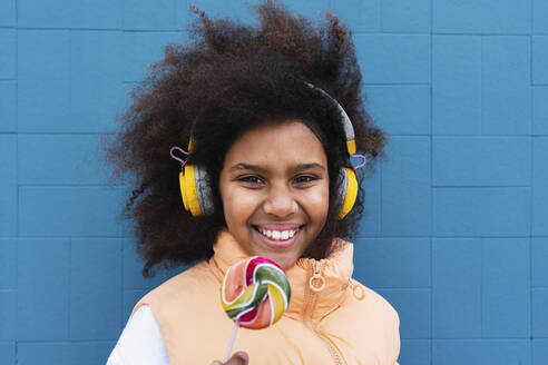 Lächelndes Mädchen mit kabellosen Kopfhörern steht mit Süßigkeiten vor einer blauen Wand - PNAF04061