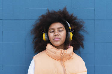 Mädchen mit geschlossenen Augen hört Musik über drahtlose Kopfhörer vor einer blauen Wand - PNAF04062