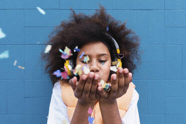 Girl wearing wireless headphones blowing confetti in front of blue wall - PNAF04063