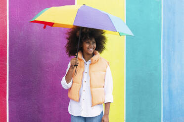 Happy girl standing with colorful umbrella in front of multi colored wall - PNAF04085