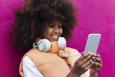 Girl wearing headphones using mobile phone leaning on pink wall - PNAF04086