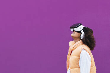 Girl wearing virtual reality simulator standing against purple background - PNAF04089