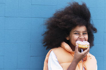 Hungry girl eating doughnut standing in front of blue wall - PNAF04090
