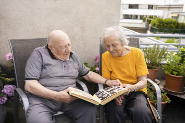 Senior man with woman looking at photo album sitting on balcony - SVKF00289