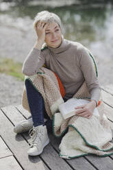 Mature woman with book and blanket sitting on pier - AANF00260