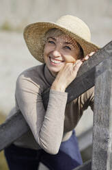 Happy mature woman wearing hat leaning on railing - AANF00263