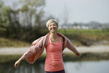 Smiling mature woman standing with arms outstretched on sunny day - AANF00289